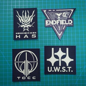 Könnte beinhalten: Vier dunkelblaue, gestickte Aufnäher mit weißen Designs und Text. Die Aufnäher enthalten die Texte "HAS", "ENDFIELD", "TCCC" und "U.W.S.T.". Die Aufnäher befinden sich auf einer grünen Gitteroberfläche.