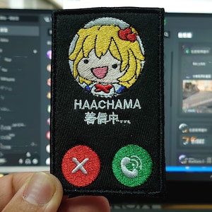 May include: A black embroidered patch featuring a cartoon character with yellow hair and a red bow. The character is smiling and has a white collar. The text "HAACHAMA 待ち中..." is below the character. The patch has a red button with a white X and a green button with a white phone icon.
