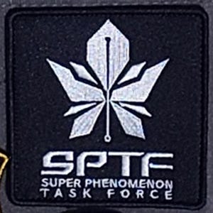 May include: Black square patch featuring a stylized white leaf design. The text "SPTF SUPER PHENOMENON TASK FORCE" is printed in white below the leaf. The patch has a textured appearance.