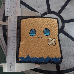 May include: Embroidered patch featuring a sad-faced character in a tan paper bag design. The character has blue eyes, a small frown, and a bandage. The patch is approximately 2.5 inches tall and 2 inches wide.