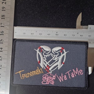 May include: A gray embroidered patch with a white heart design featuring two intertwined hands. The text "Tsunomaki" and "Wa Ta Me" is embroidered in gold and pink.