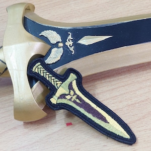 May include: A gold and black sword with a winged emblem on the blade, positioned behind a small embroidered patch shaped like a dagger. The patch features a black border, a braided handle design, and a central blade motif in shades of green and gold.
