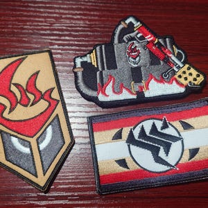 May include: Three embroidered patches featuring distinct designs: a tan shield with a red flame icon, a black and grey mechanical device with red flames, and a rectangular patch with horizontal red, white, and tan stripes and a central circular logo.