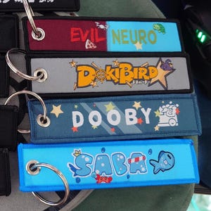 May include: A collection of embroidered keychains with various designs and text. The keychains feature the phrases "EVIL NEURO", "DokiBird", "DOOBY", and "SABA" in colorful fonts. Each keychain has a metal ring for attachment.