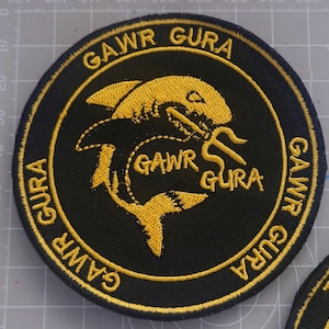 May include: A round, black embroidered patch with gold text and a gold shark illustration. The text "GAWR GURA" is repeated around the border and inside the shark design. The shark has a long, wavy tail and is facing left.