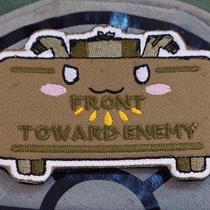 May include: Embroidered patch depicting a cartoon mine with a smiling face and the text "FRONT TOWARD ENEMY". The patch is brown with white, pink, and yellow details, set against a grey background.