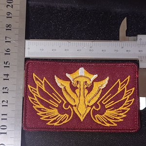 May include: A burgundy embroidered patch with a gold winged emblem. The emblem features a stylized design with a white center and gold wings.