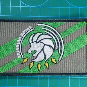 May include: Embroidered patch with a white lion-like design and the text "SHISHIRO BOTAN". The patch has a grey background with green diagonal stripes and a black border. The lion has a white mane and yellow accents.