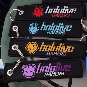 Op de afbeelding: Zwarte sleutelhangers met de tekst "hololive GAMERS" en dierlijke afbeeldingen in rood, blauw, geel en paars. Elke sleutelhanger heeft een zilveren sleutelring.