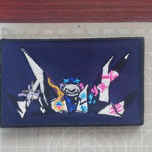 May include: Embroidered patch with a navy blue background and a colorful design. The design features a central cartoon character with a crown, surrounded by abstract shapes in white, pink, and blue. The patch is rectangular with a black border.