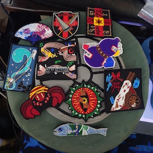 May include: A selection of embroidered patches with diverse designs. Features a fan, a shield with a sword, a cross with text, a blue and white design, a musical-themed patch, a purple bottle-shaped patch, a red and black creature, a circular design, a patch with Japanese text, and a fish.