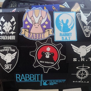 Pode incluir: Um remendo de tecido preto com vários remendos bordados anexados. Os remendos apresentam diferentes desenhos, incluindo um coelho, um urso, uma engrenagem e um floco de neve. O texto nos remendos inclui "RABBIT", "S.A.T", "RED WINTER", "S.R.T" e "S.C.H.A.L.E".