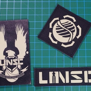 May include: Three black and white embroidered patches. One rectangular patch features the text "UNSC" and the words "United Nations Space Command" below an eagle emblem. A square patch and a rectangular patch with the text "UNSC" are also visible.