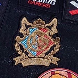 May include: Embroidered patch featuring a heraldic crest design. The crest includes a crown, shield, and laurel leaves, with blue and red accents. The patch is outlined in gold thread and has a banner at the bottom.