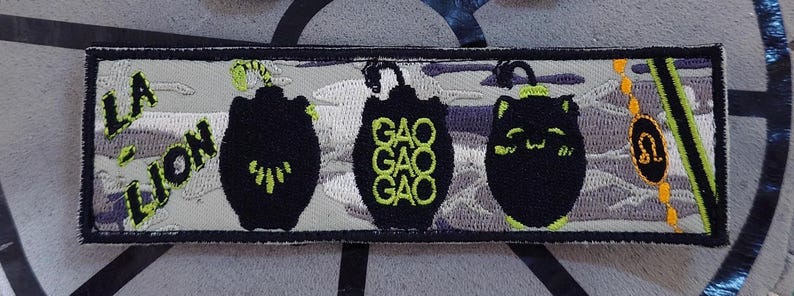 K&ouml;nnte beinhalten: Gestickter Aufn&auml;her mit schwarzem Rand, der drei stilisierte schwarze Formen mit neongr&uuml;nen Akzenten zeigt. Der Aufn&auml;her enth&auml;lt den Text "LA LION" und ein Sternzeichen-Symbol f&uuml;r L&ouml;we. Der Hintergrund hat ein wolkenartiges Muster.