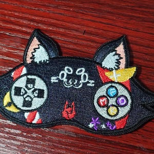 May include: A black embroidered patch shaped like a cat head with pointed ears. The design features a white directional pad on the left, a circular button array with blue, red, purple, and yellow accents on the right, and a small red icon.