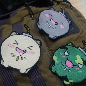 May include: Three embroidered patches featuring a cartoon bomb design. One is cream, one is grey, and one is green with a camouflage pattern. Each patch has a wick and a smiling face with pink accents.