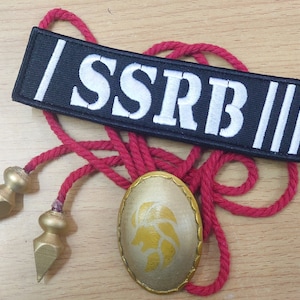 Könnte beinhalten: Eine rote Schnurkette mit zwei goldfarbenen, speerförmigen Spitzen. Die Kette hat einen goldfarbenen, ovalen Anhänger mit einem Löwenmotiv. Ein schwarzer Patch mit weißem Text, der "||SSRB||" lautet, ist ebenfalls abgebildet.