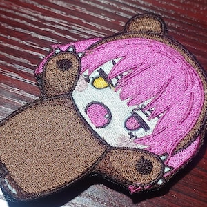 Può includere: Una toppa ricamata raffigurante un personaggio dei cartoni animati con capelli rosa acceso che indossa un costume da orso marrone. Il personaggio ha il viso bianco, occhi gialli e un'espressione sorpresa con la bocca aperta. Il costume ha orecchie rotonde.