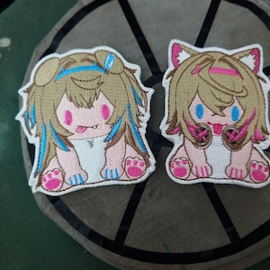 May include: Two embroidered patches featuring anime-style characters with long blonde hair, pink cheeks, and blue eyes. One character is wearing a white dress and has blue and white accents. The other character is wearing a white dress with pink and black accents. Both characters have a pink tongue sticking out.