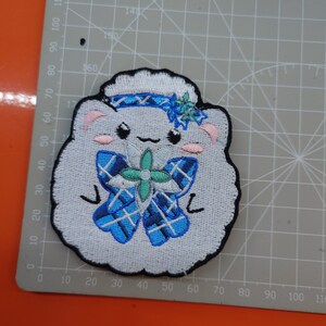 May include: White embroidered patch of a cartoon sheep wearing a blue and white plaid bow with a green flower on top. The sheep has pink cheeks and a blue headband.