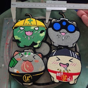 以下が含まれることがあります： 漫画の猫のデザインが施された4つの刺繍パッチ。猫はさまざまな色で、帽子やヘアバンドを着用しています。1匹の猫は黄色の帽子をかぶった緑色で、もう1匹は青い目の灰色で、他の2匹は食べ物に関連するデザインです。