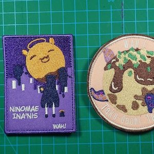 May include: Two embroidered patches. One is rectangular, purple, with a cartoon character and the text "Ninomae Ina'nis" and "WAH!". The other is round, beige, with a takoyaki design and the text "Tako dachi Tako-yaki".