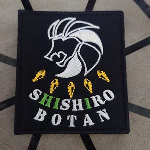 May include: Black embroidered patch featuring a white lion head design and yellow diamond shapes. The text "SHISHIRO BOTAN" is embroidered in white with green accents.