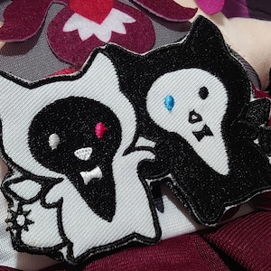 May include: Two black and white embroidered patches of cartoon ghost characters. One ghost has a red eye and the other has a blue eye.