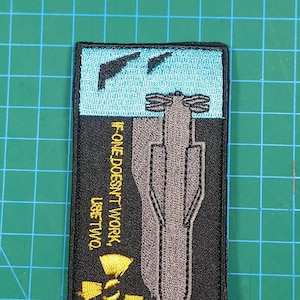 Puede incluir: Parche bordado con borde negro. El parche presenta un diseño de bomba gris, un símbolo de radiación amarillo y el texto "IF ONE DOESN'T WORK, USE TWO." La sección superior tiene un fondo azul con dos formas negras.