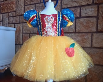 May include: A Snow White-inspired costume, featuring a yellow tulle skirt with glitter and a red apple applique. The bodice is red and blue with sequin detailing, and a matching red bow headband completes the look. The costume is displayed on a white mannequin.