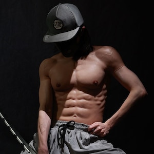 Puede incluir: Una persona con una gorra de béisbol gris con la palabra "STOP", pantalones de chándal grises y sosteniendo una espada. La persona está arrodillada sobre un fondo oscuro.