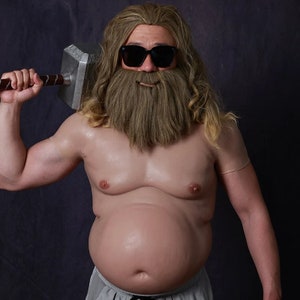 Puede incluir: Un hombre con una peluca larga rubia, gafas de sol y una barba postiza sostiene un martillo plateado. Lleva un body de color carne y pantalones cortos grises.