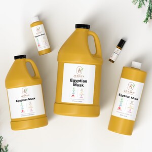 May include: Four bottles of Egyptian Musk essential oil in different sizes. The bottles are yellow and have a white label with the text "Jersey Botanicals Egyptian Musk" on them. The bottles are arranged on a white surface with green foliage in the background.
