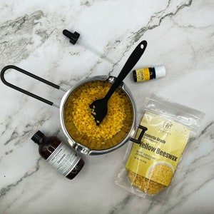 May include: A stainless steel double boiler with a black silicone spatula stirring yellow beeswax pellets. A bag of yellow beeswax pellets labeled "Cosmetic Grade Triple Filtered Yellow Beeswax" is on a white marble surface. A small bottle of jasmine essential oil and a bottle of lemon essential oil are also on the surface.