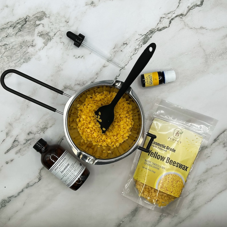 May include: A stainless steel double boiler with a black silicone spatula stirring yellow beeswax pellets. A bag of yellow beeswax pellets labeled "Cosmetic Grade Triple Filtered Yellow Beeswax" is on a white marble surface. A small bottle of jasmine essential oil and a bottle of lemon essential oil are also on the surface.