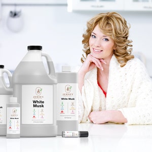 May include: Four bottles of White Musk essential oil in various sizes, from a small roller bottle to a large 32 oz bottle. The bottles are all white with a black label and the Jersey Botanicals logo. The bottles are sitting on a white table in front of a woman wearing a white sweater.