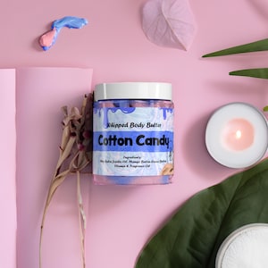 May include: A jar of "Cotton Candy" whipped body butter with a white lid and blue accents. The jar is surrounded by pink and green leaves, a lit candle, and a pink background. The ingredients are listed on the jar.
