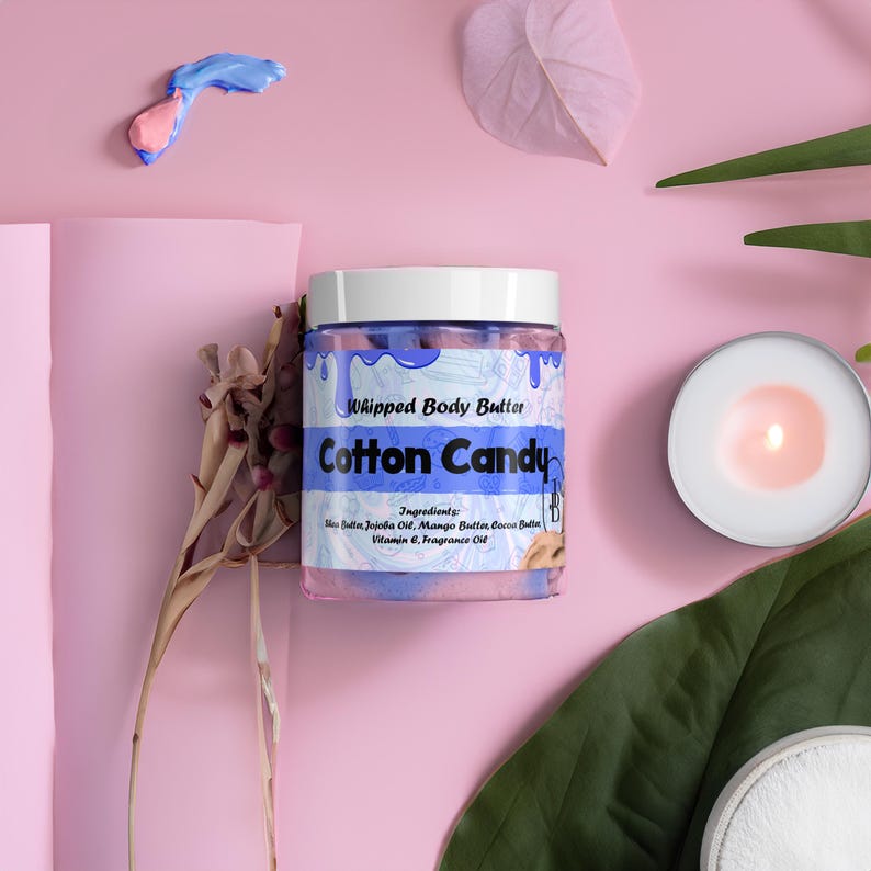 May include: A jar of "Cotton Candy" whipped body butter with a white lid and blue accents. The jar is surrounded by pink and green leaves, a lit candle, and a pink background. The ingredients are listed on the jar.