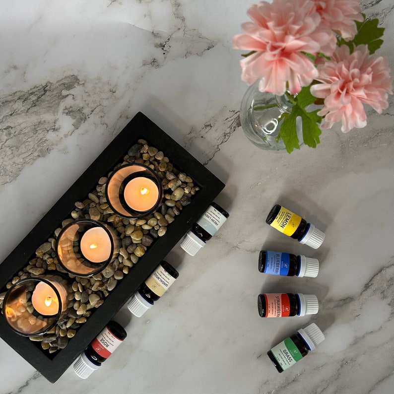 May include: A black wooden tray with three glass candle holders filled with lit candles and small stones. Six essential oil bottles are arranged on a white marble surface. The bottles are labeled with the names of the oils: Lemon, Lavender, Tea Tree, Frankincense, Rose, and Grass.