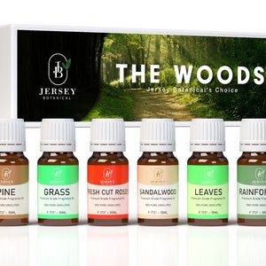 May include: A set of six 10 ml bottles of essential oils with woodsy scents. The bottles are labeled with the names "Pine", "Grass", "Fresh Cut Roses", "Sandalwood", "Leaves", and "Rainforest". The bottles are arranged in a row on a white surface. The background is a blurred image of a forest path.