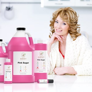 May include: Four bottles of pink sugar scented liquid in various sizes, with the Jersey Botanicals logo and product name on the labels. The bottles are arranged on a white surface with a woman in the background.
