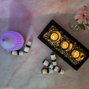 May include: A black tray with three lit candles surrounded by small stones.  There are seven small bottles of essential oils on a pink marble surface next to the tray.  A white diffuser with a purple light is also on the surface.