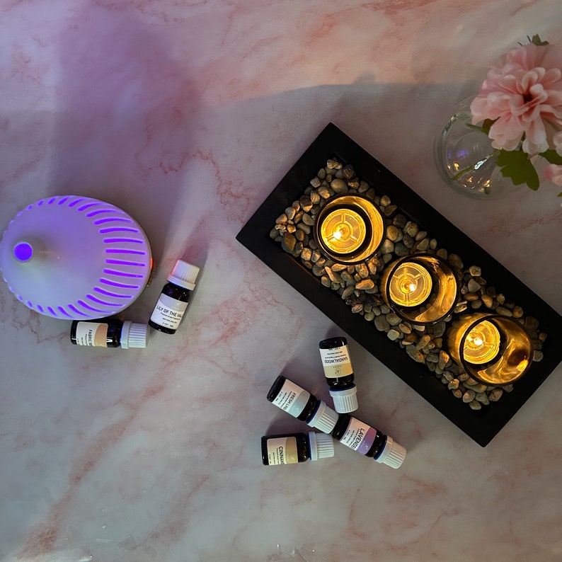 May include: A black tray with three lit candles surrounded by small stones.  There are seven small bottles of essential oils on a pink marble surface next to the tray.  A white diffuser with a purple light is also on the surface.