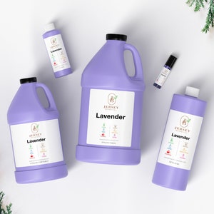 Op de afbeelding: Vier flessen lavendelolie in verschillende maten. De flessen zijn paars en hebben een wit etiket met de tekst "Jersey Botanicals Lavender". De flessen zijn gerangschikt op een wit oppervlak met een takje groen blad op de achtergrond.