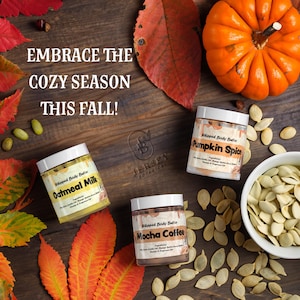 May include: An autumnal arrangement featuring three jars of whipped body butter with labels reading "Oatmeal Milk", "Pumpkin Spice", and "Mocha Coffee". A bright orange pumpkin, fall leaves, and pumpkin seeds are scattered around the jars. The text "EMBRACE THE COZY SEASON THIS FALL!" is visible.