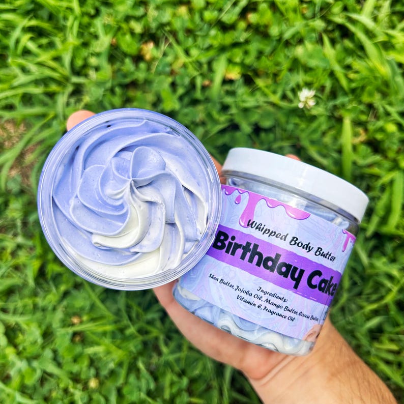 May include: A jar of "Whipped Body Butter" with "Birthday Cake" text, held in a hand. The jar is clear with a white lid and a label with purple and pink accents. The body butter inside is swirled with blue and white colors.