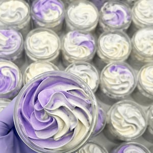 May include: A collection of small, clear jars filled with whipped body butter. The body butter is swirled with white and lavender colors, creating a decorative pattern. The jars are arranged in rows, with one jar held in a gloved hand.