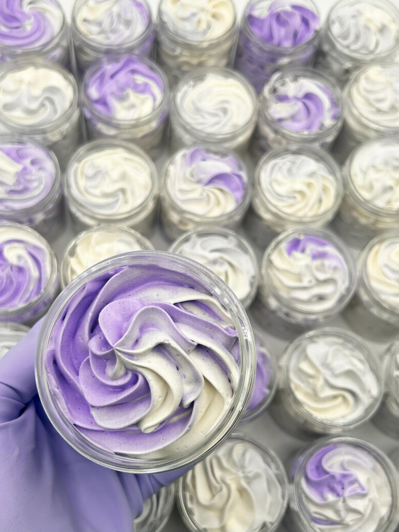 May include: A collection of small, clear jars filled with whipped body butter. The body butter is swirled with white and lavender colors, creating a decorative pattern. The jars are arranged in rows, with one jar held in a gloved hand.