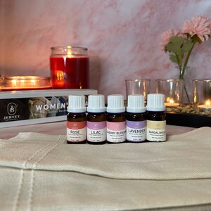 May include: Five small glass bottles with white lids and labels. The bottles are filled with essential oils and labeled with the scents: Rose, Lilac, Cherry Blossom, Lavender, and Sandalwood. The bottles are arranged in a row on a white surface. The text "WOMEN'S" is visible in the background.